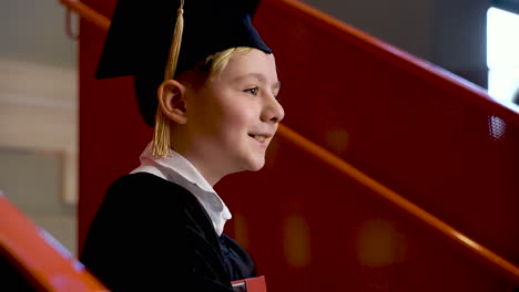 side view of a happy preschool male student in cap and gown holding book at the graduation ceremony