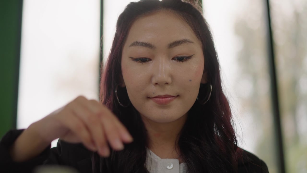 Primo piano di una giovane donna con lunghi capelli scuri, che indossa grandi orecchini a cerchio e abiti casual, mantenendo un'espressione calma. Una luce naturale soffusa le illumina il viso.