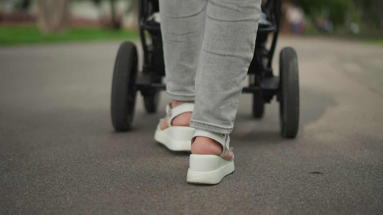 Closeup of feet walking stroller, gray jeans and white sandals on asphalt path, rhythmic step and spinning wheels, low camera perspective, urban park routine, active parental lifestyle influenced