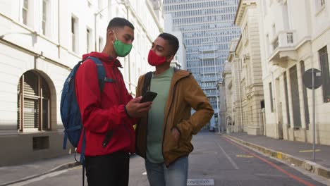 two happy mixed race male friends standing, talking and using smartphone in the street