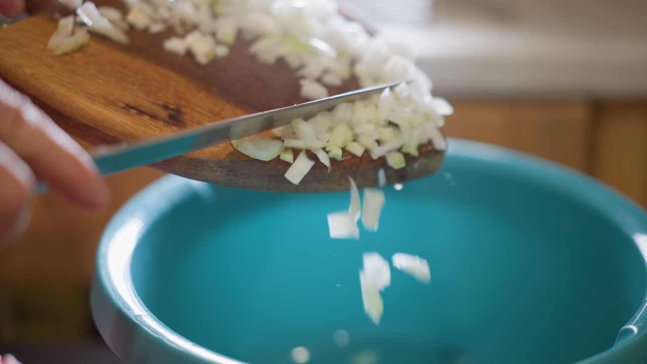 Closeup of chopping onion. Preparing kitchen with onion scraps into container in natural light. Closeup of person transferring onion pieces from cutting board to bowl under natural lighting