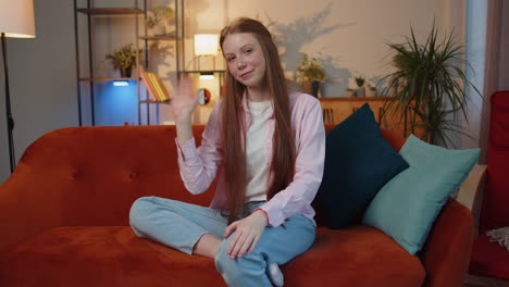 child girl smiling friendly at camera, waving hands gesturing hello, hi, greeting at home on couch