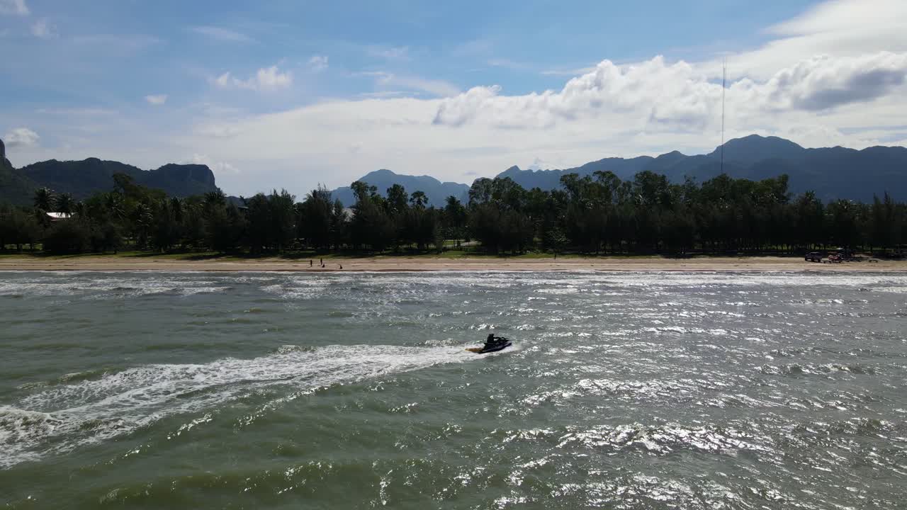 una silueta de moto acuática acelerando hacia la derecha en diagonal a la orilla, frente a la playa, montañas al fondo, tarde calurosa, bahía de los delfines, parque nacional sam roi yot, prachuap khiri khan, tailandia