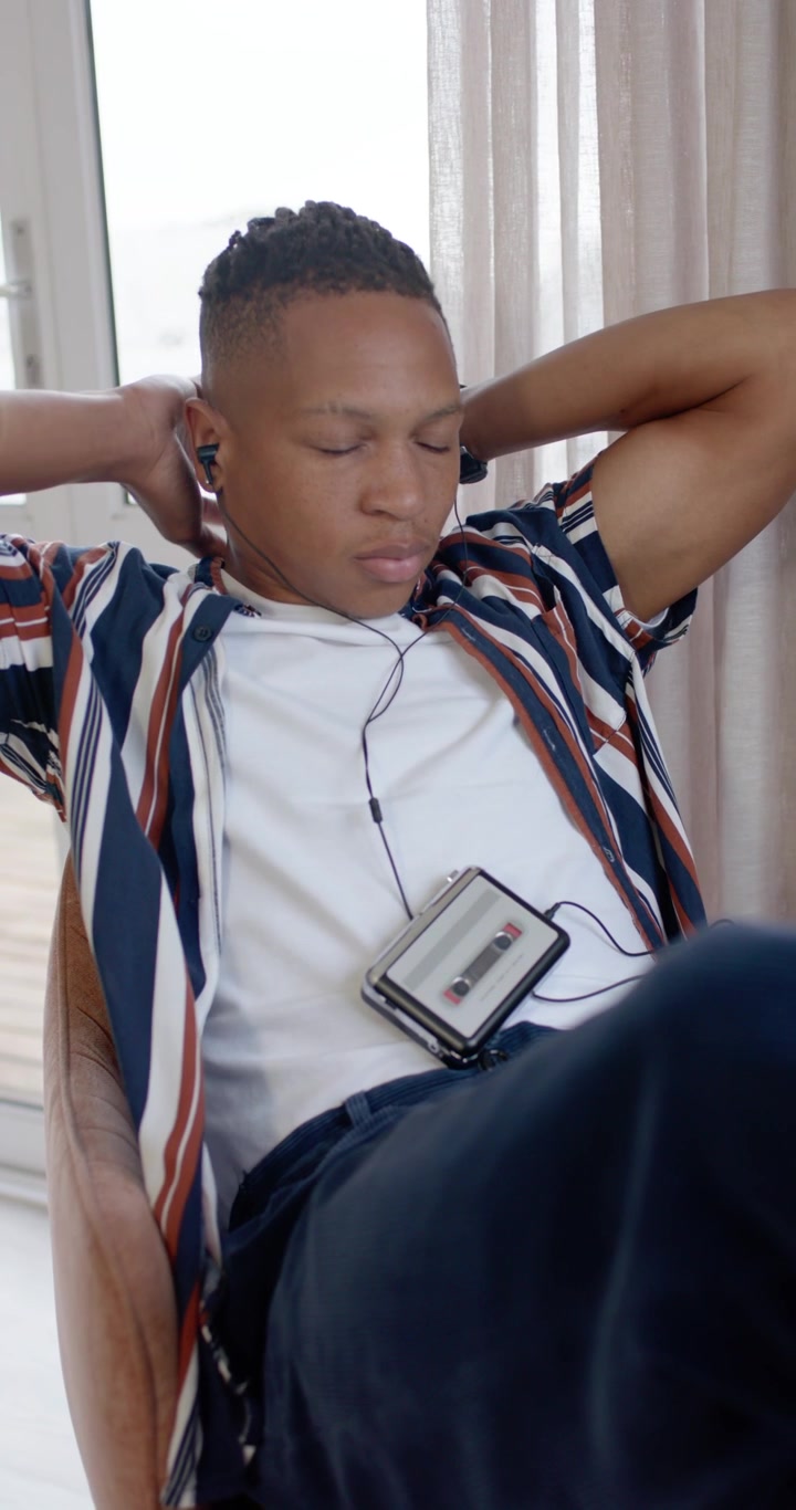 Vertical video: Adult man holding Walkman and wired earphones at window, leaning back to relax