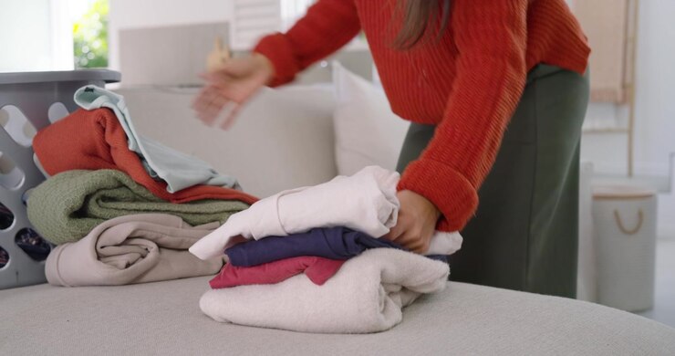 Woman is entering frame from right, folding towels and stacking clothes on ottoman to tidy room