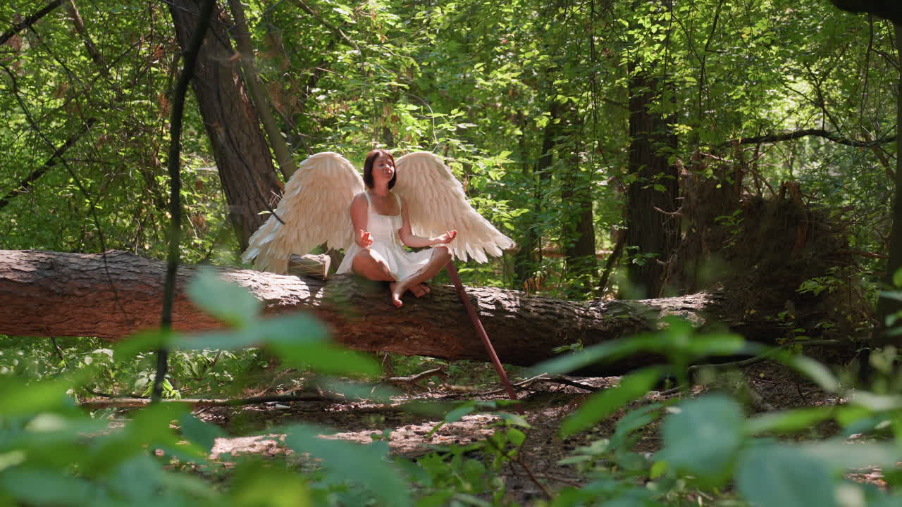 Guardiana della foresta con grandi ali d'angelo in abito bianco seduta a gambe incrociate su un tronco nella foresta mentre pratica yoga, circondata dalla luce del sole e da una vegetazione lussureggiante, con un tamburo e un bastone di legno appoggiati accanto ai suoi piedi