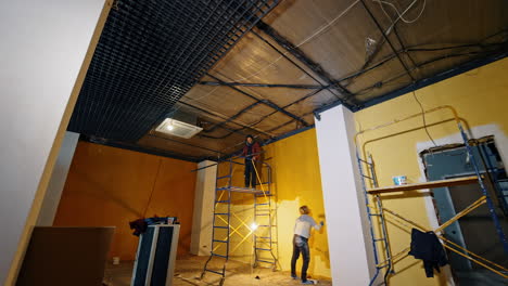builders perform repair and finishing works in the newly-built house. man installs the equipment and decor on the ceiling and woman paints the walls yellow.