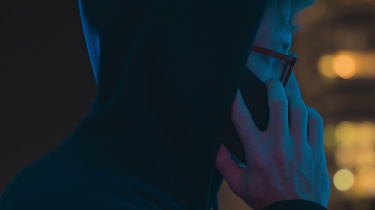 Chiamata segreta. Uomo incappucciato che effettua una telefonata segreta di notte con le luci della città sullo sfondo. Comunicazione anonima, cybercrimine, sorveglianza, spionaggio e attività criminale. Un hacker che comunica