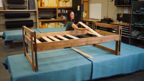 a young woman smiles enjoying herself as she builds an entire bed frame one piece of oak wood at a time in a professional carpentry workshop