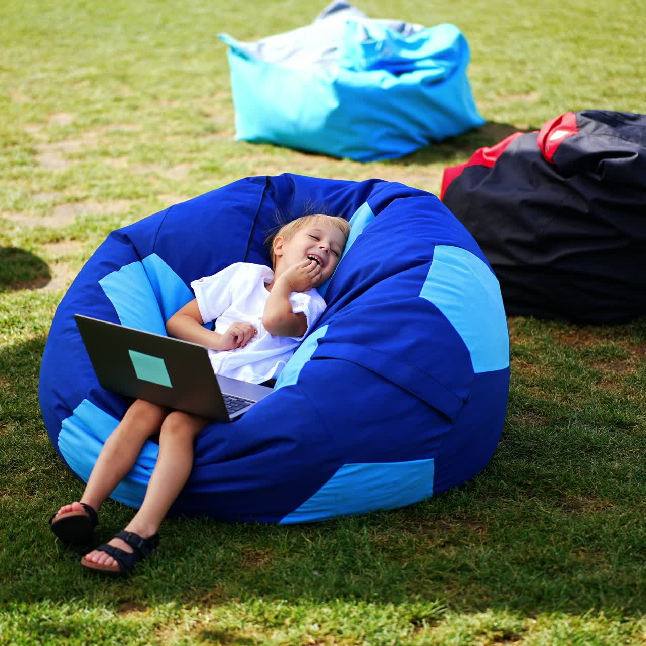 Cute boy in white shirt sits in bean bag chair with laptop on his knees. Kid watched video and laughs leaning back in chair