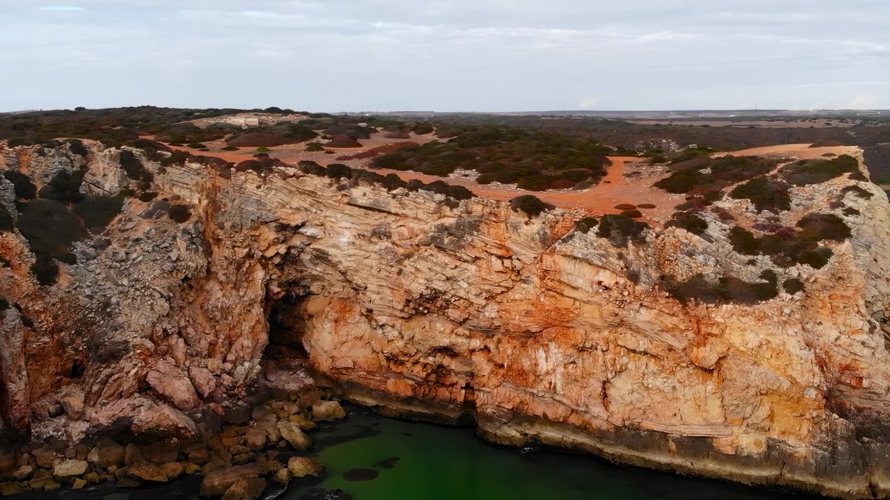 Costa a scogliera rossa frastagliata vista dall'alto, con terreno ocra e pareti calcaree esposte, arbusti sparsi e vista panoramica