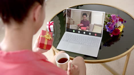 Caucasian-woman-holding-red-gift-using-laptop-with-african-american-man-on-screen