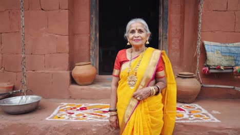 portrait of a smiling senior woman wearing a yellow sari and gold jewelry standing in front of her rural house entrance decorated with rangoli and terracotta pots
