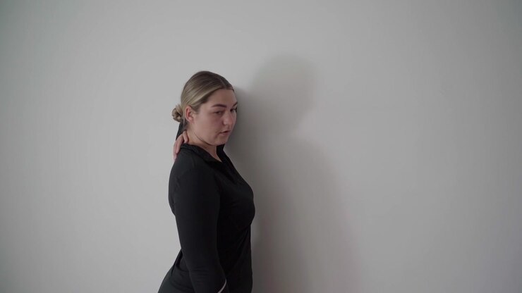 Una donna esegue uno stretching in piedi per spalle e petto contro una parete bianca, usando la posizione delle braccia per aprire la parte anteriore del corpo e alleviare la tensione legata alla scrivania