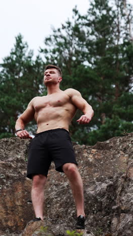 athletic man with naked torso wearing black shorts stands on the rock. sportsman rises hands showing muscles. low angle view. vertical video