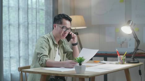 asian man designer talking on smartphone while looking at the layout bond in hand at the office