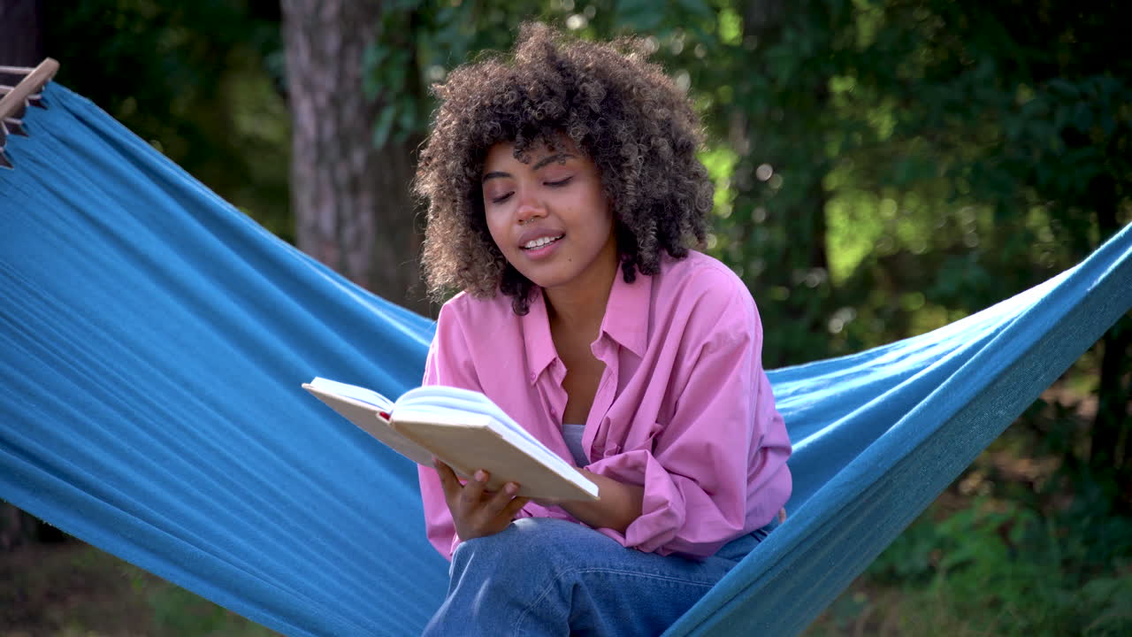 Young black woman reads a book sitting in a hammock in the woods. Pretty female with afro hair.