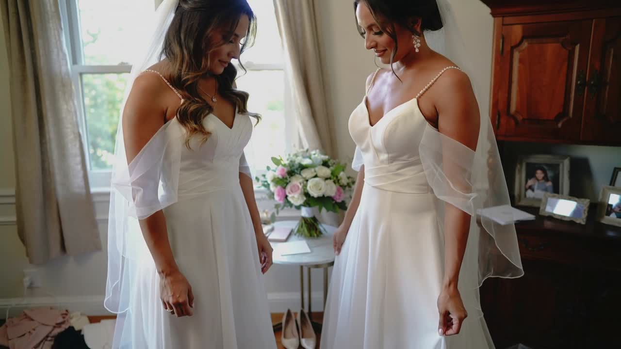 Deux mariées en robes et voiles lèvent les yeux et se tendent les mains dans une chambre, se préparant pour leur mariage.