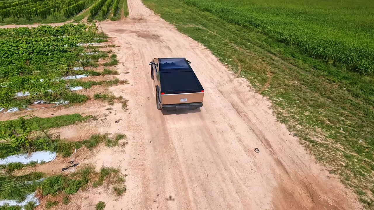 Modern Tesla Cybertruck riding quickly by the ground road. FPV drone follows the futuristic vehicle in the farmland.