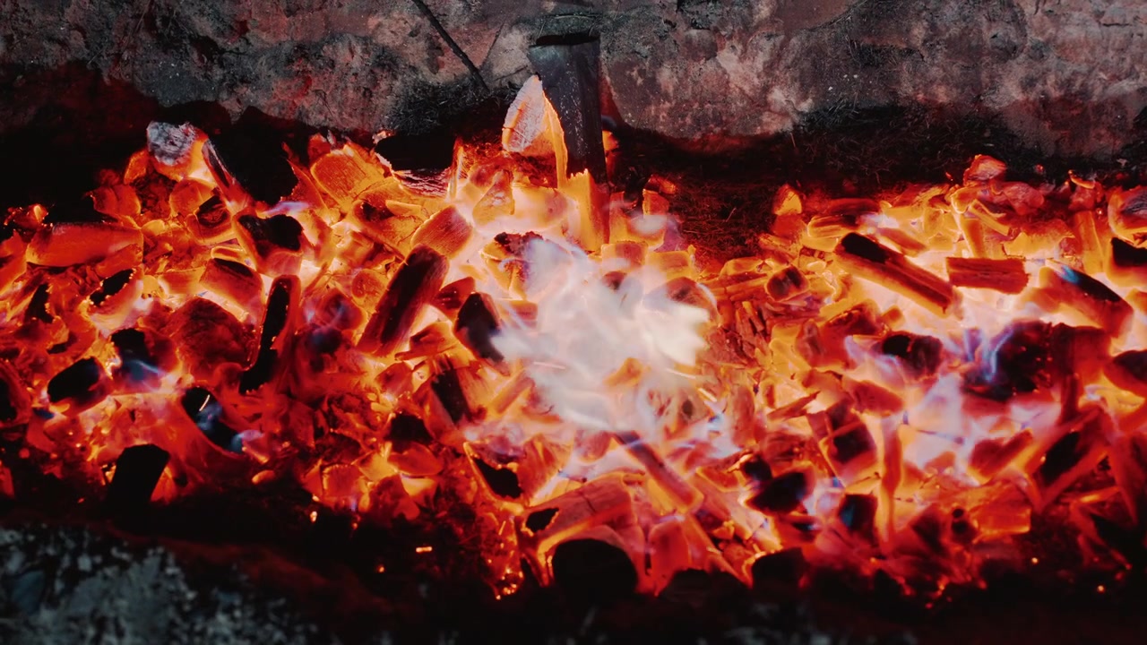 Vista ravvicinata dall'alto di un braciere rettangolare pieno di carbonella incandescente e braci ardenti, mentre fiamme vigorose si alzano e tremolano sui carboni crepitanti, proiettando una luce calda e intensa, con brace e fiamma.
