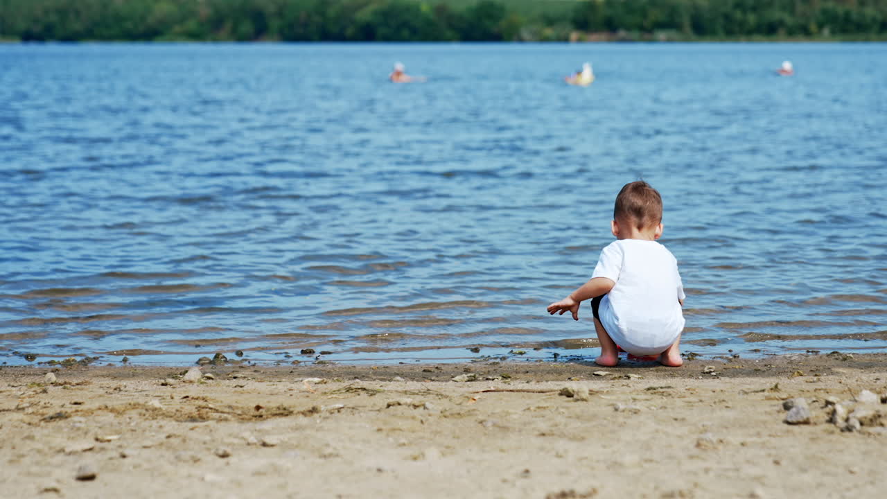 Lovely kid in white T-shirt comes up to a river. Toddler is filling his watering can with water.