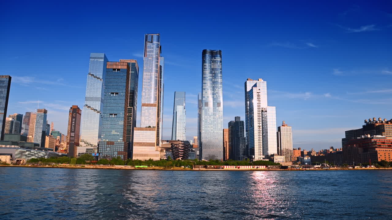 New York, USA, 1 August 2025: Sun reflects in the high-rise façade and on the riverscape. Low angle view at the waterfront of New York with Vessel Structure among the high-rises