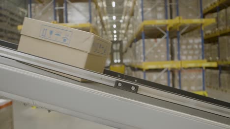 A-cardboard-box-moves-on-a-conveyor-belt-in-a-large-warehouse-with-stacked-shelves-in-the-background