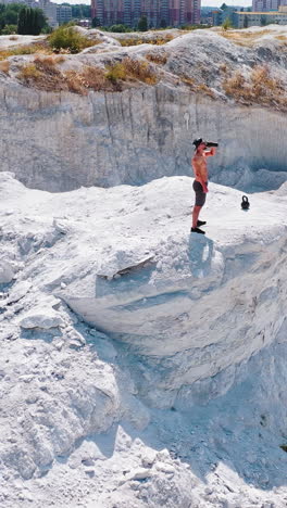 sportsman drinks water on a white canyon. strong man standing on the edge of a hill near the city. aerial view. camera moves round. vertical video