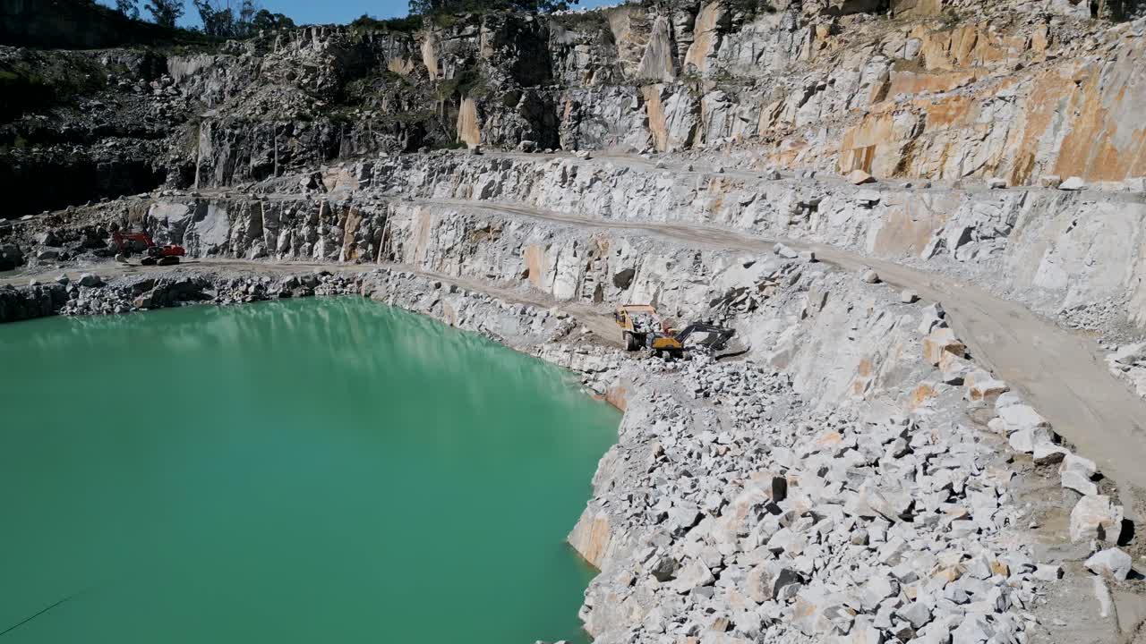 Excavator loading stone into a truck beside a turquoise quarry lake - aerial