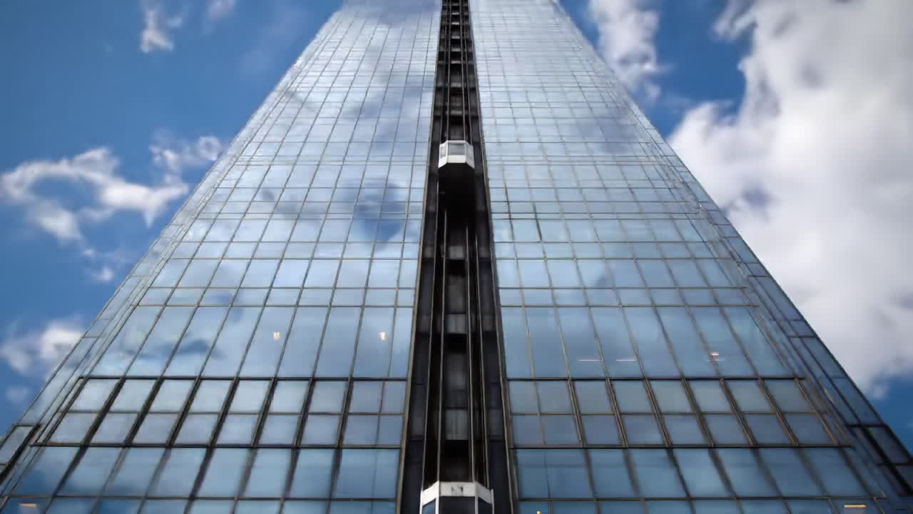 A striking view of a modern skyscraper showcasing its sleek glass exterior and unique elevator design against a backdrop of a bright, cloudy sky