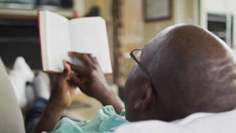 Blick-über-Die-Schulter-Eines-älteren-Afroamerikanischen-Mannes,-Der-Entspannt-Auf-Der-Couch-Liegt-Und-Ein-Buch-Liest