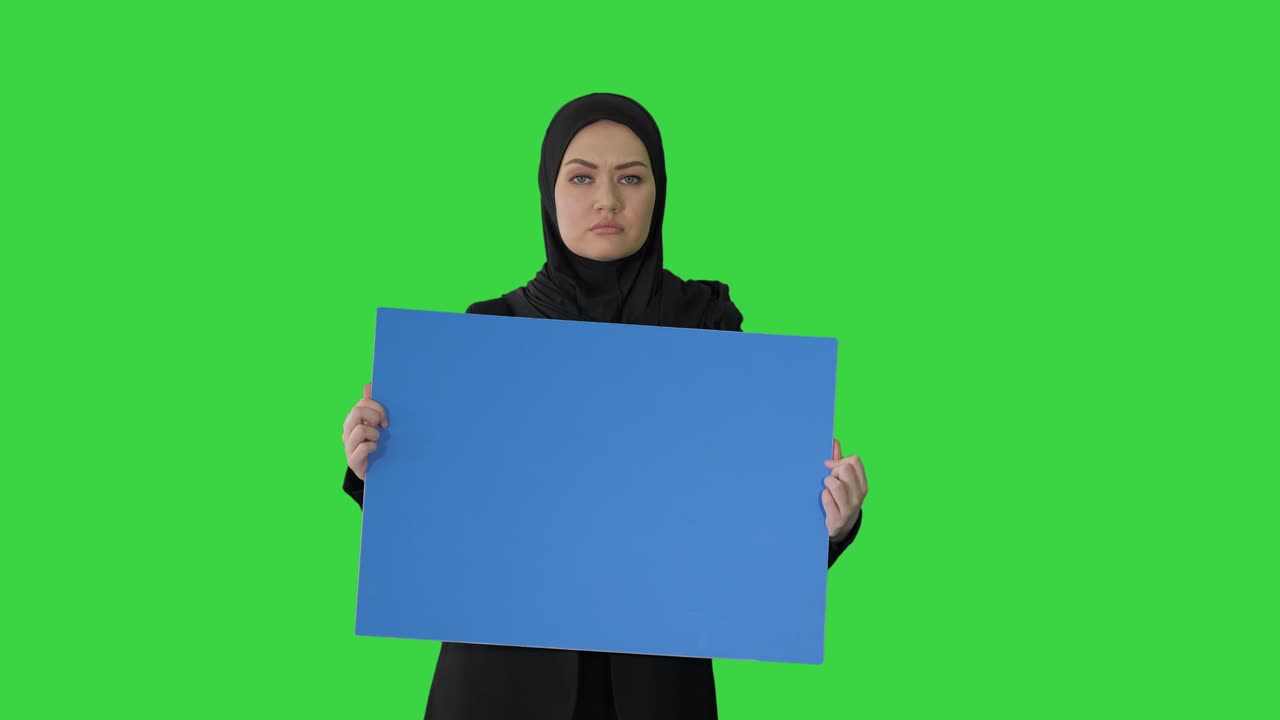 mujer árabe seria en hijab sosteniendo un cartel azul en blanco y mirando a la cámara en una pantalla verde, llave de croma