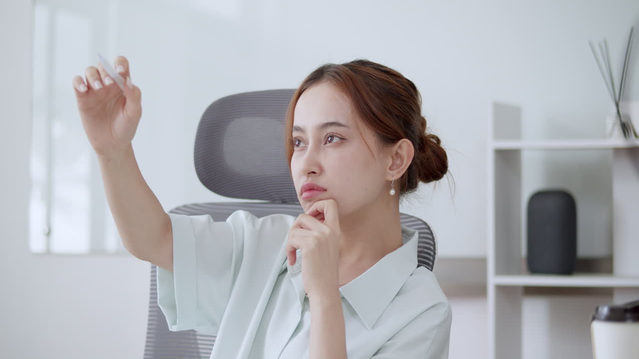A focused young woman brainstorming at her desk, using expressive gestures with a pen to spark new ideas and tap into creative inspiration.
