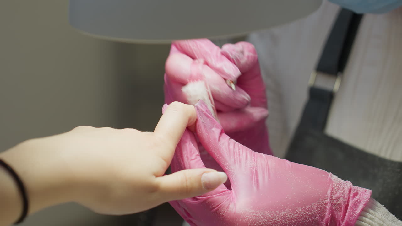 Nail technician wearing pink gloves using brush and electric filer on customer's fingernails during manicure session, with customer wearing black bracelet and technician in protective apron and mask