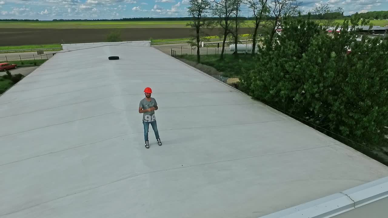 Worker on a flat roof of the industrial building. Man in safety helmet stands on roof and looking at camera. Aerial view.