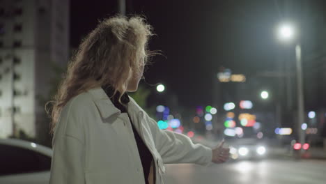 woman extends hand to stop cab while walking away from parked white car under bright streetlights in vibrant nighttime city environment filled with bokeh lights and moving traffic