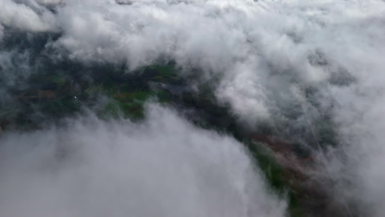 High altitude aerial view flying forward over white clouds with patches of green fields and land in Latvia visible below