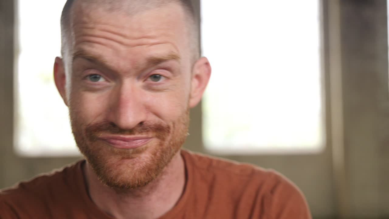 Video of a man with large rose tinted sparkly glasses laughing and talking at the camera in an industrial warehouse.