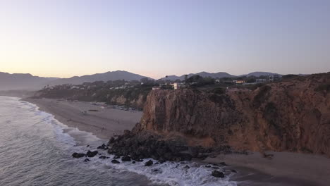 Hermoso-Acantilado-Rocoso-De-La-Costa-En-California-Al-Atardecer,-Carro-Aéreo-A-La-Izquierda-Con-Un-Hermoso-Paisaje-Montañoso-En-El-Fondo