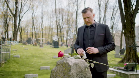 hombre de traje negro frente a una lápida con rosa roja mientras se seca las lágrimas y luego sale del cementerio