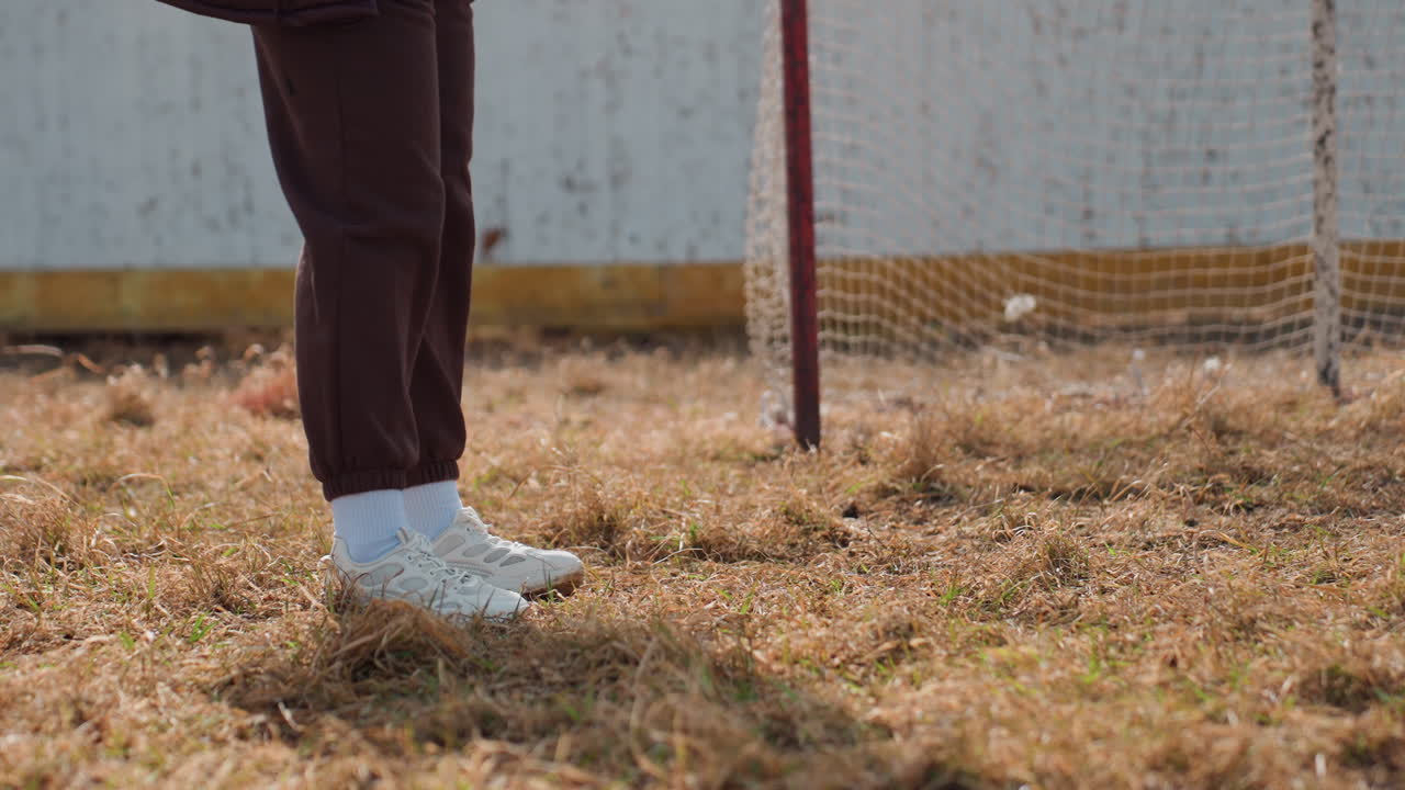 젊은 선수가 고르지 못한 경기장에서 가만히 서 있어요. 빈티지 축구 골대 근처에서 캐주얼한 옷을 입고 있는 한 사람이 야외에서 낡은 축구 골대 옆에서 조용히 멈춰 서 있네요.