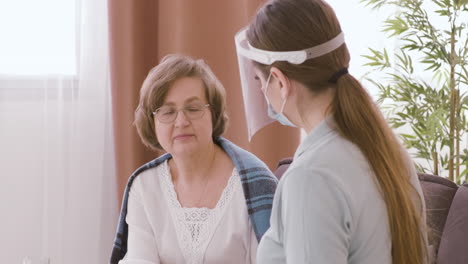 Doctora-Con-Máscara-Facial-Y-Pantalla-Protectora-Sentada-Con-Una-Anciana-Paciente-Mirando-La-Cámara