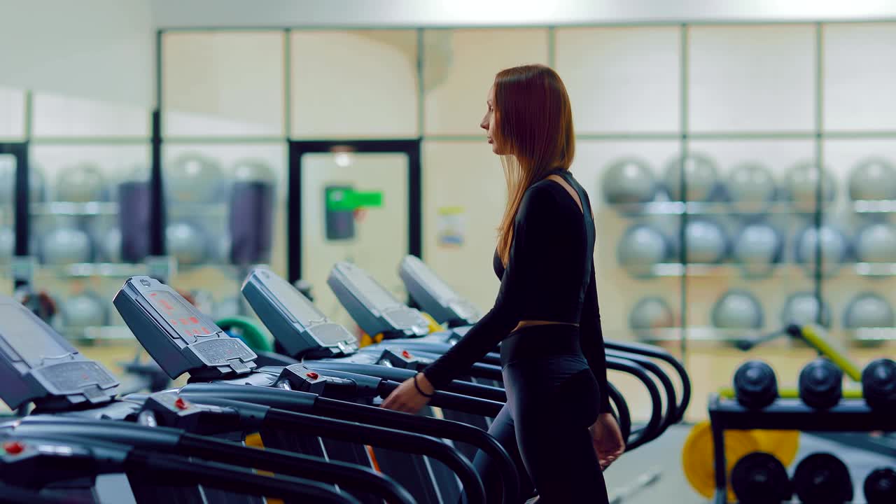 Young pretty woman is training in the gym, jogging machines are standing in a row. Pretty woman is walking on a treadmill, warming up.