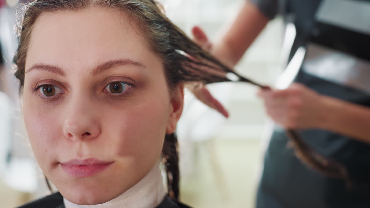 미용실에서 머리 시술과 섹션을 받는 클라이언트의 클로즈업 인물 사진, 젖은 머리카락과 차분한 표정으로 뒤에서 작업하는 스타일리스트, 은은한 주변광과 전문적인 망토, 평온한 미용 트리트먼트