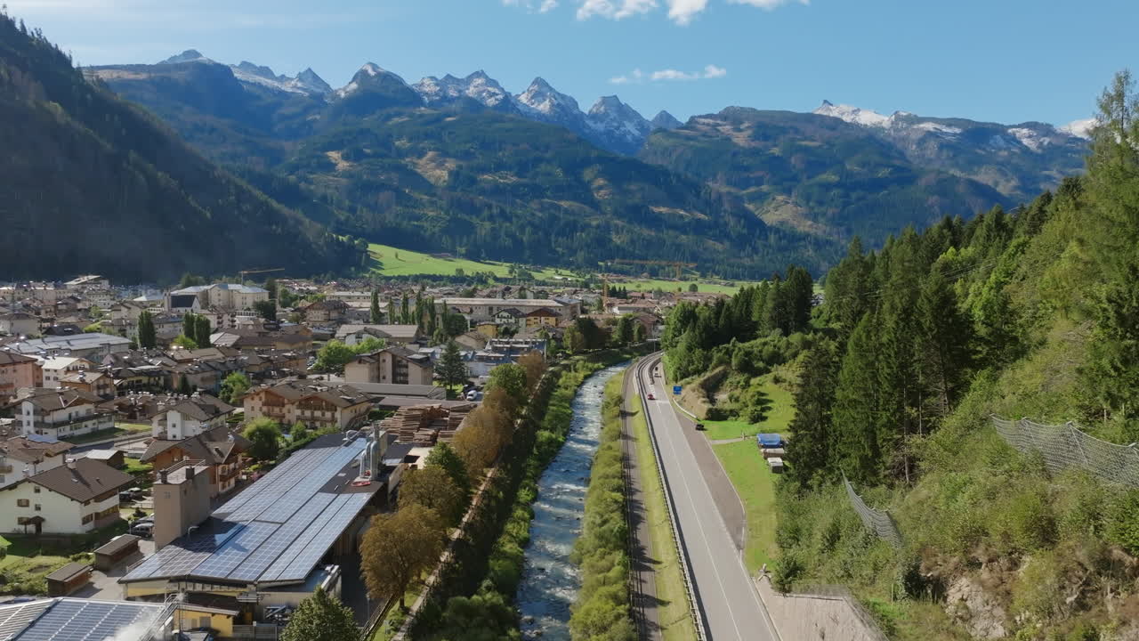 Aerial footage flying over a river, road, and Italian town in northern Italy with the Dolomite Mountains in the background.