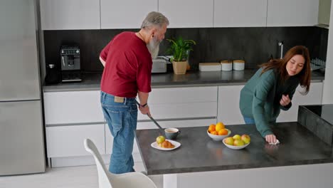 An-elderly-man-with-gray-hair-and-a-beard-in-a-red-T-shirt-is-cleaning-the-kitchen-with-a-mop-along-with-his-adult-brunette-daughter-in-a-green-sweater-in-a-modern-kitchen