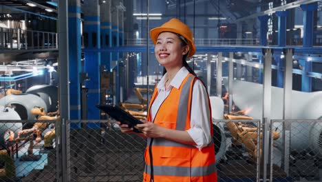 vista lateral de una ingeniera asiática con casco de seguridad mirando la tableta en su mano y mirando a su alrededor mientras está de pie en la fábrica de fabricación de turbinas eólicas