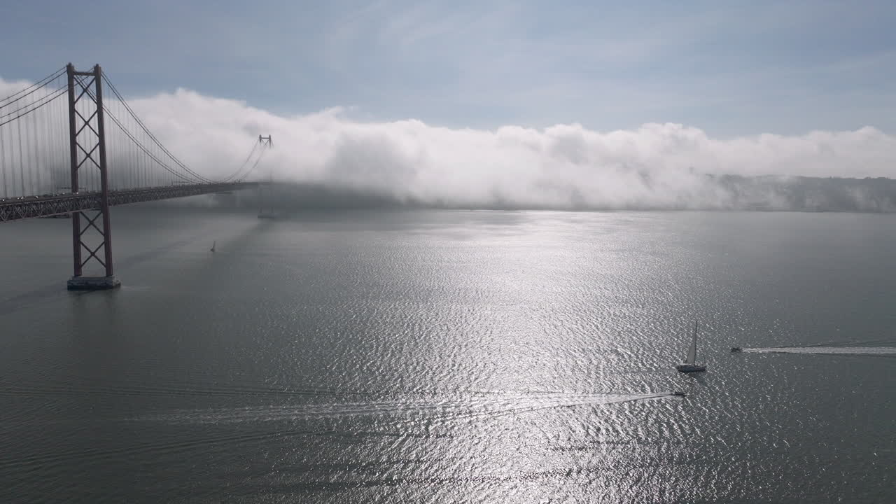 Cinematic aerial drone establishing shot towards the 25th April Bridge and waterfront in Lisbon, Portugal, Europe. Sunny and bright with clouds, fog, mist covering the bridge
