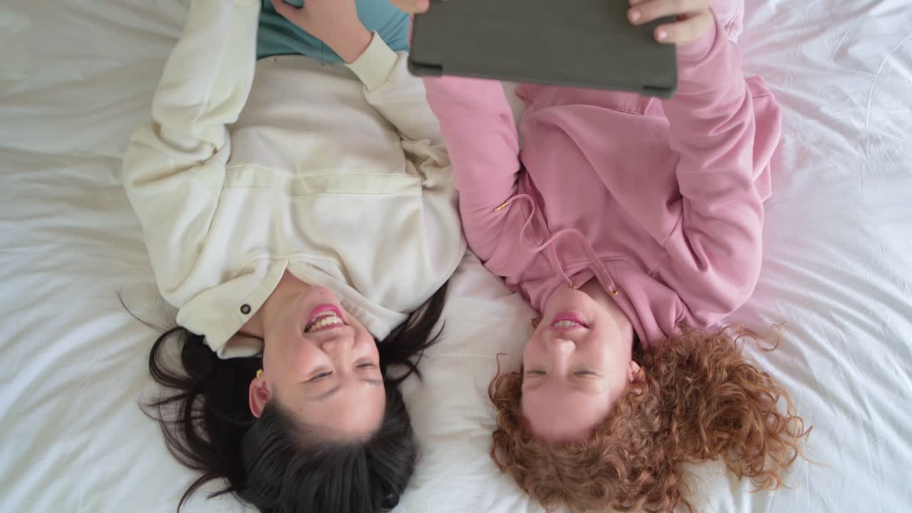 Amigas asiáticas e ruivas, a ruiva segurando um tablet, compartilhando clip engraçado enquanto estão deitadas na cama