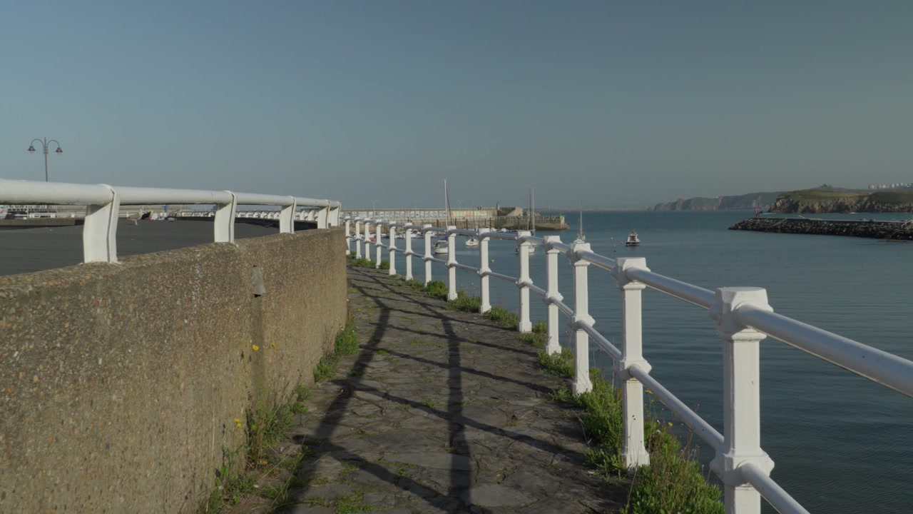 Coastal walkway with white railings following the harbor in Candás, Asturias, Spain. Scenic seaside path overlooking calm waters of the Cantabrian Sea with a peaceful maritime atmosphere.